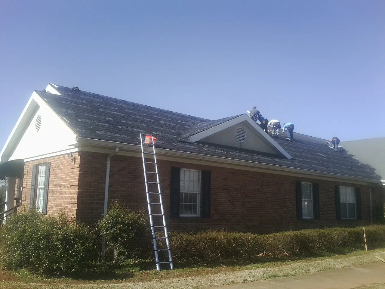 Crew tearing shingles off a brick ranch roof with a ladder set — Metal Roof Repair work by our crew in Lockport